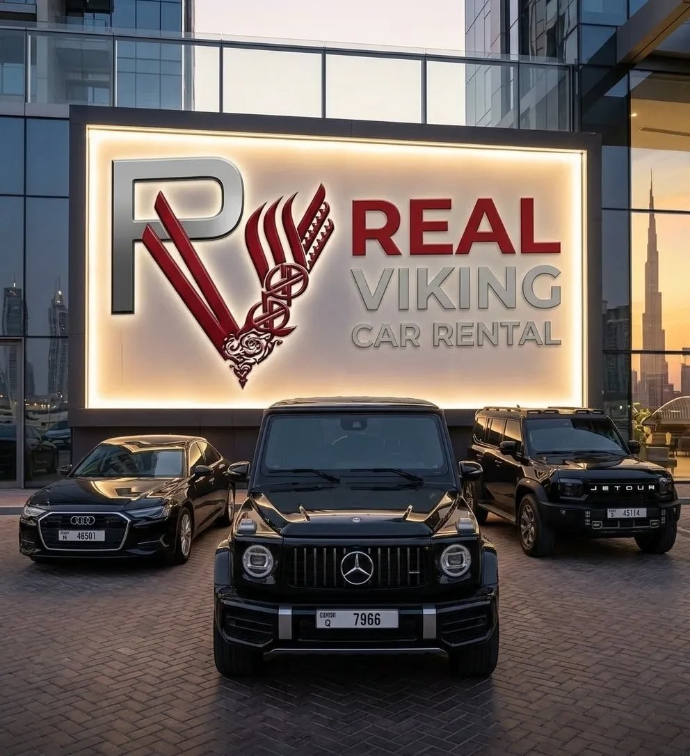 The illuminated Real Viking Car Rental sign at the Dubai showroom at dusk, with a Mercedes G-Class, an Audi A6 and a Jetour SUV parked in front.